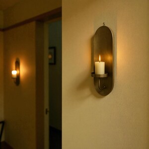 May include: A wall-mounted candle holder with a lit white candle. The holder is made of a bronze-colored metal and has an oval shape. The candle holder is mounted on a beige wall, and another candle holder is visible in the background.