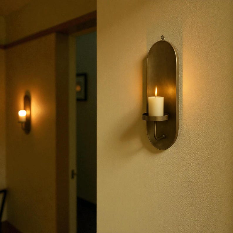 May include: A wall-mounted candle holder with a lit white candle. The holder is made of a bronze-colored metal and has an oval shape. The candle holder is mounted on a beige wall, and another candle holder is visible in the background.