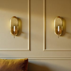 May include: Two gold-colored wall sconces with an oval shape and textured surface. Each sconce holds a white candle. The sconces are mounted on a beige wall with decorative molding.