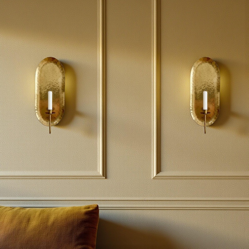 May include: Two gold-colored wall sconces with an oval shape and textured surface. Each sconce holds a white candle. The sconces are mounted on a beige wall with decorative molding.