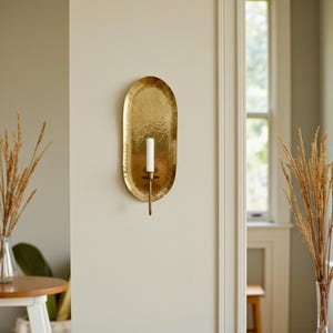 May include: A gold-colored, oval-shaped wall sconce with a hammered texture. It features a single white candle and a long, slender holder. The sconce is mounted on a neutral-colored wall, adding a touch of elegance to the space.