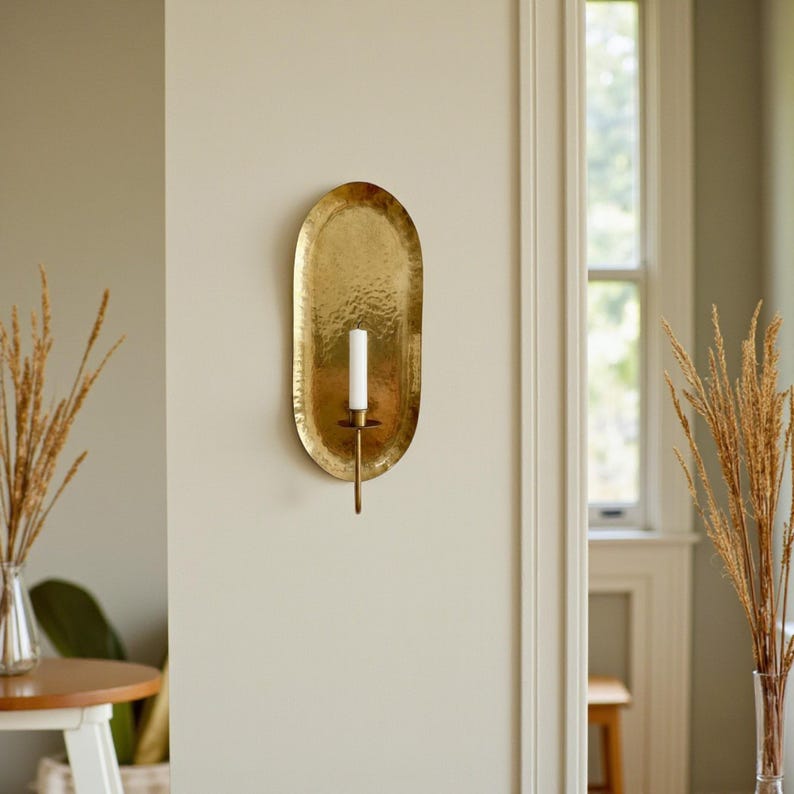 May include: A gold-colored, oval-shaped wall sconce with a hammered texture. It features a single white candle and a long, slender holder. The sconce is mounted on a neutral-colored wall, adding a touch of elegance to the space.