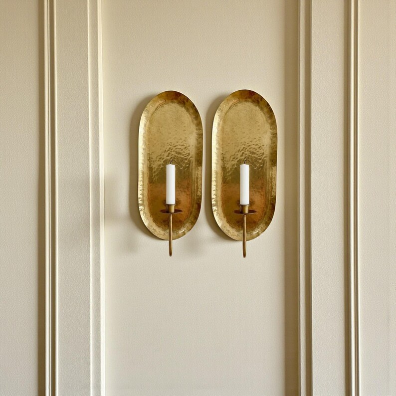 May include: Two gold-colored, oval wall sconces with a hammered texture. Each sconce holds a white candle. The sconces are mounted on a cream-colored wall with vertical trim.