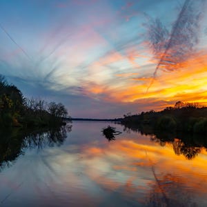 Puede incluir: Fotografía de un paisaje de un lago al atardecer. El cielo transita del azul al naranja y amarillo vibrantes, reflejándose en el agua en calma. Los árboles bordean las orillas, agregando profundidad a la escena tranquila.