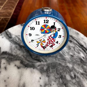 May include: A vintage blue alarm clock with a white face featuring a colorful clown illustration. The clock has a silver top and a white border around the face. The clock face displays black numbers and red star markers.