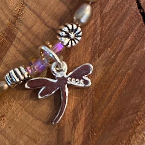 May include: Close-up of a beaded bracelet with a dragonfly charm. The bracelet features gold-colored beads, silver accents, and a dragonfly charm with the marking "S925". The dragonfly charm is silver with a dark red outline. The background is a wooden surface.
