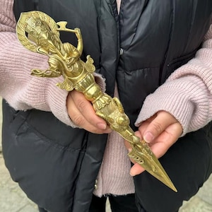 May include: A detailed brass ritual dagger, held in hands. The dagger features intricate carvings of a deity and ornate patterns. The handle has a figure with a halo, and the blade is pointed. The overall design is golden and detailed.
