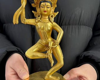 May include: A gold-coloured metal statue of a deity in a dancing pose, featuring an ornate crown, a third eye on the forehead, and jewellery. The figure stands on a circular base with a lotus petal design, holding a small object in the raised right hand.