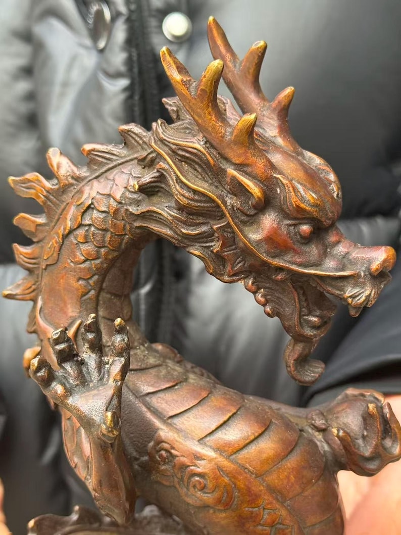 K&ouml;nnte beinhalten: Eine bronzefarbene Drachenskulptur mit aufwendigen Details. Der Drache hat H&ouml;rner, Schuppen und eine geschwungene Haltung. Die Skulptur ist in Bronze- und Braunt&ouml;nen gehalten und zeigt das detaillierte Design der Kreatur.