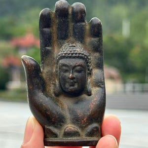 May include: A bronze-colored sculpture of Buddha's head within an open hand. The sculpture has intricate details, with a serene expression on the face. The background is blurred, suggesting an outdoor setting.