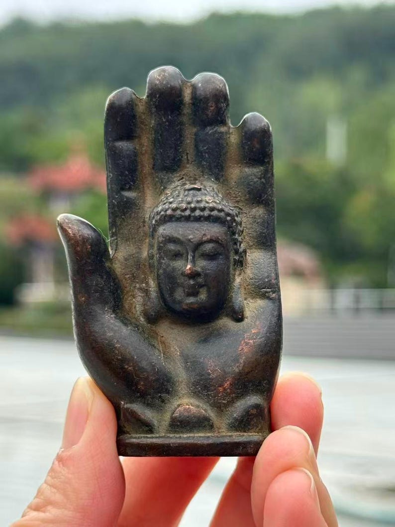 May include: A bronze-colored sculpture of Buddha's head within an open hand. The sculpture has intricate details, with a serene expression on the face. The background is blurred, suggesting an outdoor setting.