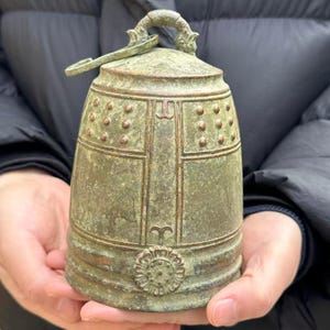 Pode incluir: Um sino de cor bronze com uma alça de laço e um badalo. O sino tem um design detalhado com um padrão floral na base e uma série de pequenos detalhes redondos. O sino é segurado nas mãos de uma pessoa.