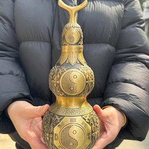 Puede incluir: Un objeto decorativo en forma de doble calabaza, de color latón. El objeto presenta intrincados patrones florales y geométricos, junto con tres símbolos de Yin y Yang. La parte superior tiene un asa estilizada y bifurcada. El objeto se sostiene frente a una persona que lleva una chaqueta negra.