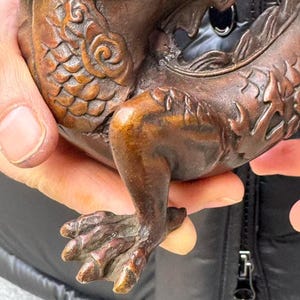 K&ouml;nnte beinhalten: Nahaufnahme einer Bronzeskulptur, die eine mythische Kreatur, m&ouml;glicherweise einen Drachen, darstellt. Die Skulptur zeigt detaillierte Handwerkskunst mit Schuppen, Krallen und Zierornamenten. Die Bronze hat eine reiche, gealterte Patina mit verschiedenen Braun- und Kupfert&ouml;nen.