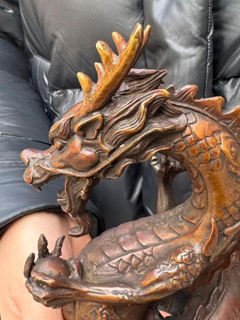K&ouml;nnte beinhalten: Bronzefarbene Drachenskulptur mit aufwendigen Details. Der Drache hat einen detaillierten Kopf mit Geweih, offenem Maul und Schuppen. Die Skulptur ist ein Dekorationsartikel, wahrscheinlich zur Pr&auml;sentation.