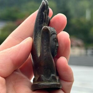 May include: Bronze-colored sculpture of a Buddha head held within a hand. The sculpture has detailed facial features and is mounted on a small base. The hand is also bronze-colored and appears to be holding the Buddha head.