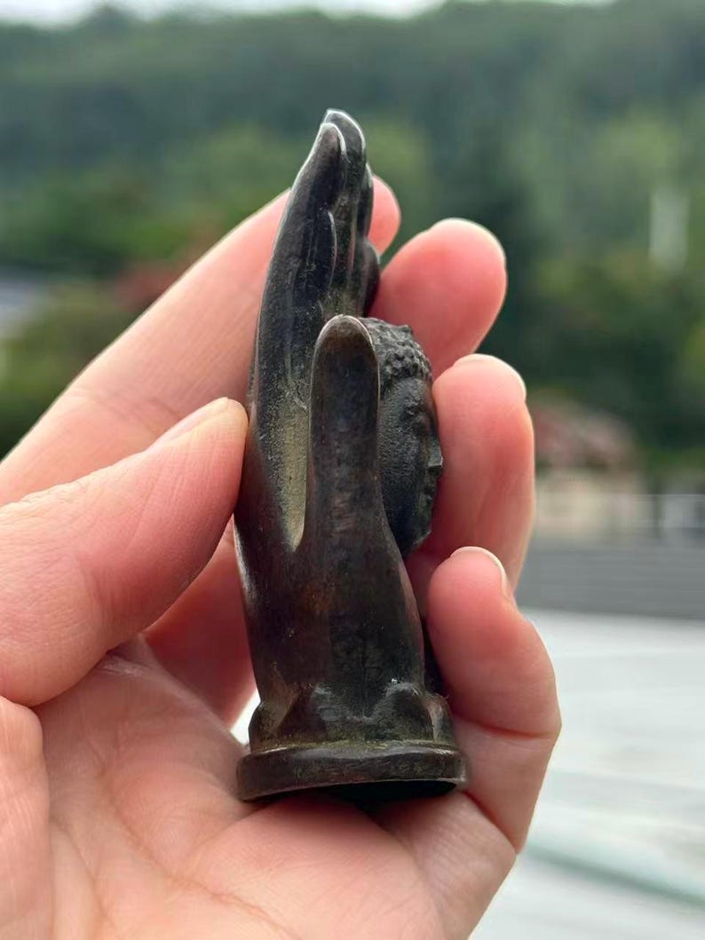 May include: Bronze-colored sculpture of a Buddha head held within a hand. The sculpture has detailed facial features and is mounted on a small base. The hand is also bronze-colored and appears to be holding the Buddha head.
