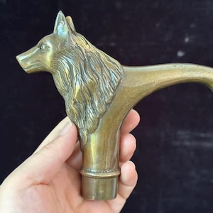 Könnte beinhalten: Ein Gehstockgriff aus Messing in Form eines Wolfskopfes mit Hals. Die Skulptur zeigt detaillierte Fellstrukturen, spitze Ohren und einen geschwungenen Hals, der in einen Griff übergeht, mit einem zylindrischen Sockel zur Befestigung am Stock.