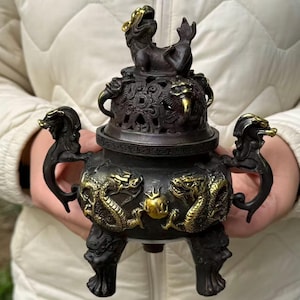 May include: A bronze incense burner with intricate dragon and mythical creature detailing. The burner features a lid with a small statue and handles with dragon heads. The base has three feet and is decorated with gold accents.