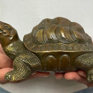 May include: A bronze turtle figurine with detailed shell and textured skin. The sculpture is held in hands, showcasing its antique finish and intricate design. The turtle's head is raised, and its legs are extended, giving it a lifelike appearance.