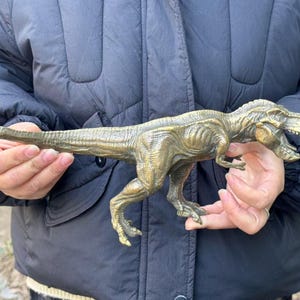 May include: A metallic bronze-colored Tyrannosaurus Rex figurine featuring a textured, scaly surface, a long tail, sharp claws, and an open mouth showing teeth. The dinosaur is depicted in a standing position with its head turned to the side.