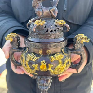 May include: A bronze incense burner with gold accents. The burner features dragon and mythical creature designs. The lid is topped with a mythical creature. The handles are dragon-shaped. The burner is held by a person.