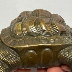 May include: A detailed bronze turtle figurine. The turtle's shell has a textured, layered design, and the body is intricately detailed. The figurine is a warm, antique gold color, suggesting it is made of metal. The turtle's head and legs are visible.