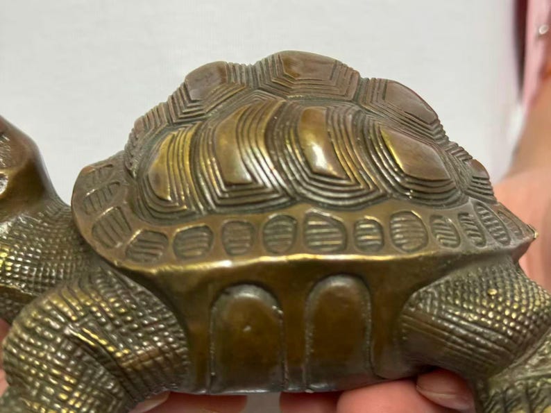 May include: A detailed bronze turtle figurine. The turtle's shell has a textured, layered design, and the body is intricately detailed. The figurine is a warm, antique gold color, suggesting it is made of metal. The turtle's head and legs are visible.