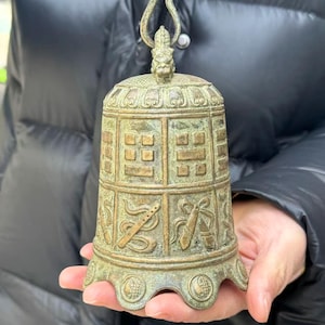 May include: Bronze bell with intricate relief carvings. The bell features a handle with a dragon-like figure and detailed patterns around the body. The bell is held in a person's hands.