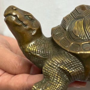 May include: A bronze-colored turtle figurine with detailed texture. The turtle's head is raised, and its shell features a geometric pattern. The figurine is held in a person's hands, showcasing its size and craftsmanship.