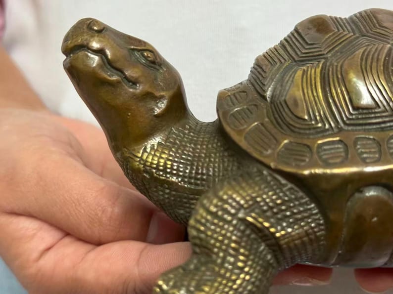 May include: A bronze-colored turtle figurine with detailed texture. The turtle's head is raised, and its shell features a geometric pattern. The figurine is held in a person's hands, showcasing its size and craftsmanship.