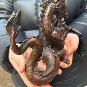 K&ouml;nnte beinhalten: Eine detaillierte Bronzedrachenskulptur. Der Drache ist in einer geschwungenen Pose dargestellt, wobei sein Schwanz und Kopf einen Kreis bilden. Die Skulptur hat eine strukturierte Oberfl&auml;che und eine satte, braune Farbe, die die Schuppen, H&ouml;rner und Krallen hervorhebt.