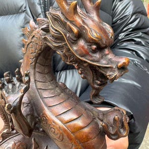 K&ouml;nnte beinhalten: Bronzene Drachenskulptur mit aufwendigen Details. Der Drache hat ein detailliertes Gesicht mit H&ouml;rnern, eine lange Schnauze und eine flie&szlig;ende M&auml;hne. Der K&ouml;rper ist mit Schuppen bedeckt, und die Krallen sind sichtbar. Die Skulptur hat eine satte Bronzefarbe.