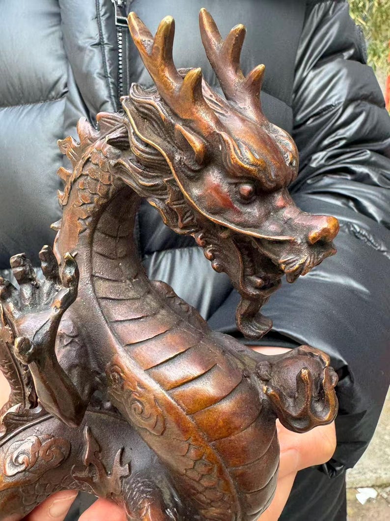 K&ouml;nnte beinhalten: Bronzene Drachenskulptur mit aufwendigen Details. Der Drache hat ein detailliertes Gesicht mit H&ouml;rnern, eine lange Schnauze und eine flie&szlig;ende M&auml;hne. Der K&ouml;rper ist mit Schuppen bedeckt, und die Krallen sind sichtbar. Die Skulptur hat eine satte Bronzefarbe.