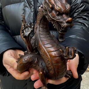 K&ouml;nnte beinhalten: Bronzene Drachenskulptur mit aufwendigen Details. Der Drache ist in einer dynamischen Pose dargestellt, mit ausgestreckten Krallen und erhobenem Kopf. Die Skulptur hat eine dunkle, braune Patina, die die Schuppen und Merkmale hervorhebt.