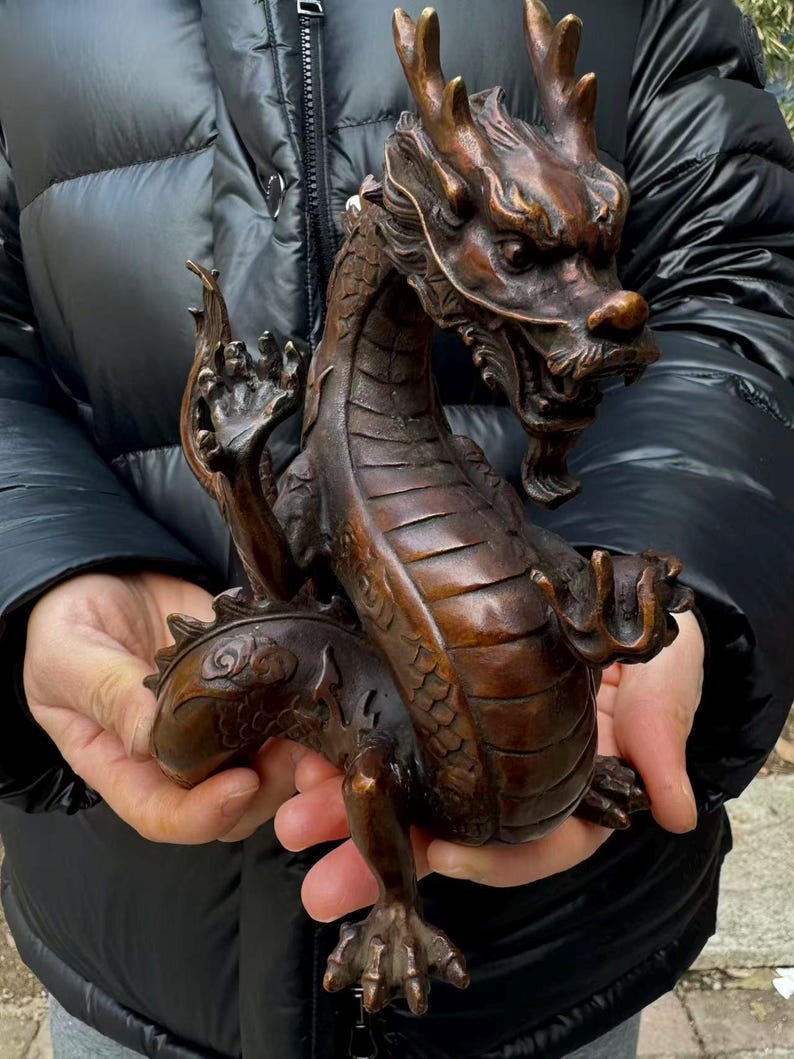 K&ouml;nnte beinhalten: Bronzene Drachenskulptur mit aufwendigen Details. Der Drache ist in einer dynamischen Pose dargestellt, mit ausgestreckten Krallen und erhobenem Kopf. Die Skulptur hat eine dunkle, braune Patina, die die Schuppen und Merkmale hervorhebt.