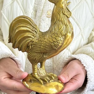 May include: A gold-colored metal rooster figurine standing on a circular base. The sculpture features detailed feather textures on the wings, tail, and neck, with a prominent comb and wattle on the head. The rooster is depicted in a standing pose.