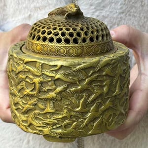 May include: A detailed, antique brass incense burner. The lid features a honeycomb pattern and a small bird-like sculpture. The body is adorned with a raised relief design. The overall color is a warm, aged gold.