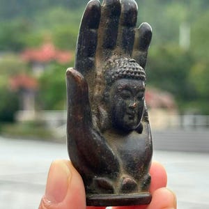 May include: A bronze-colored sculpture of a Buddha head nestled within a hand. The sculpture has intricate details, with the Buddha's face and the hand's fingers clearly defined. The background is blurred, suggesting an outdoor setting.