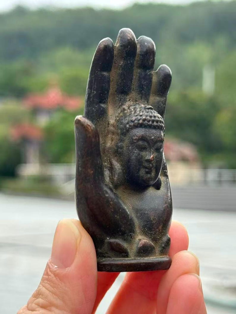 May include: A bronze-colored sculpture of a Buddha head nestled within a hand. The sculpture has intricate details, with the Buddha's face and the hand's fingers clearly defined. The background is blurred, suggesting an outdoor setting.