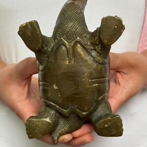 May include: A bronze-colored turtle figurine with its legs and head extended. The turtle's shell has a textured pattern, and the figurine appears to be made of metal. The turtle is positioned upside down, showcasing its underside.