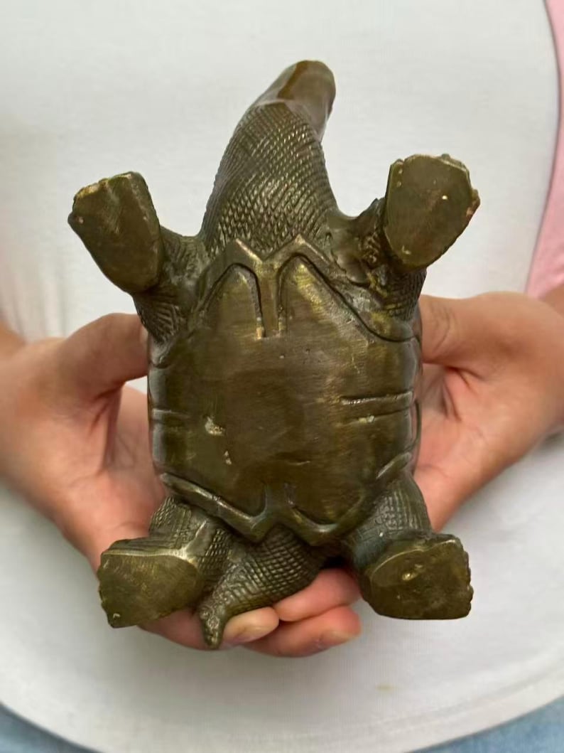 May include: A bronze-colored turtle figurine with its legs and head extended. The turtle's shell has a textured pattern, and the figurine appears to be made of metal. The turtle is positioned upside down, showcasing its underside.