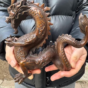 K&ouml;nnte beinhalten: Eine Bronzedrachenskulptur mit detaillierten Schuppen, H&ouml;rnern und einem flie&szlig;enden Schwanz. Der Drache ist gebogen und wird von H&auml;nden gehalten. Die Skulptur hat ein verwittertes, antikes Aussehen.