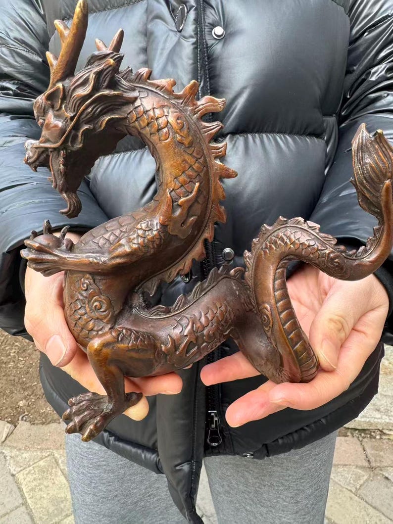 K&ouml;nnte beinhalten: Eine Bronzedrachenskulptur mit detaillierten Schuppen, H&ouml;rnern und einem flie&szlig;enden Schwanz. Der Drache ist gebogen und wird von H&auml;nden gehalten. Die Skulptur hat ein verwittertes, antikes Aussehen.