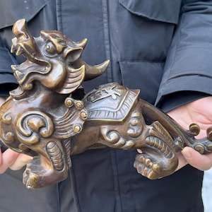 May include: A bronze-coloured metal sculpture of a mythical creature, likely a qilin or lion-dog, featuring intricate swirling patterns, a saddle-like detail on its back, an open mouth with visible teeth, and a long, curved tail extending outwards.