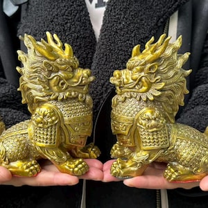 Może przedstawiać: Wyświetlane są dwa złote, bogato zdobione posągi mitycznych stworzeń, prawdopodobnie smoków lub lwów. Posągi są szczegółowe z teksturowanymi łuskami, wyszukanymi grzywami i dzikimi wyrazami twarzy. Postacie są w pozycji siedzącej.