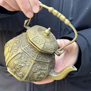 May include: A detailed, antique brass teapot with intricate relief carvings of dragons and floral patterns. The teapot has a curved spout and a handle, and a lid with a small knob. The overall design is ornate and decorative.