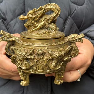 May include: A detailed brass incense burner with a dragon motif. The lid is topped with a dragon sculpture, and the body features dragon designs and ornate detailing. The burner has three feet and handles with dragon heads.