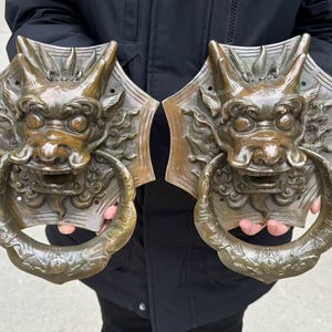 May include: Two bronze dragon door knockers. Each knocker features a detailed dragon head with an open mouth and a circular handle. The dragon heads are mounted on a shield-shaped backplate. The finish has a weathered appearance.