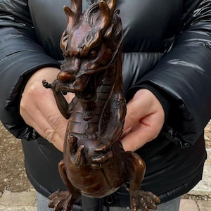 K&ouml;nnte beinhalten: Bronze-Drachenfigur mit aufwendigen Details. Der Drache hat ein detailliertes Gesicht, Geweihe und Schuppen. Er ist mit verschr&auml;nkten Armen und F&uuml;&szlig;en auf dem Boden posiert. Die Figur ist ein dekorativer Gegenstand.